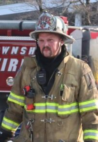 A firefighter in full gear walks on a snowy lawn holding an ax and a tool, with a fire truck and other firefighters standing in the background near a house.