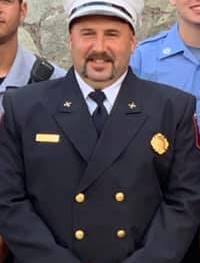 A man in a dark navy uniform with gold buttons and a badge, wearing a white cap, standing in front of a stone wall. Two other individuals, partially visible, stand beside him in blue uniforms.