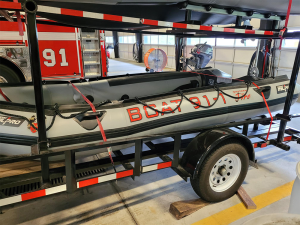 Inflatable rescue boat labeled "BOAT 91-1" is secured on a trailer inside a fire station. Red ropes hold it in place, and a fire truck is partially visible in the background.