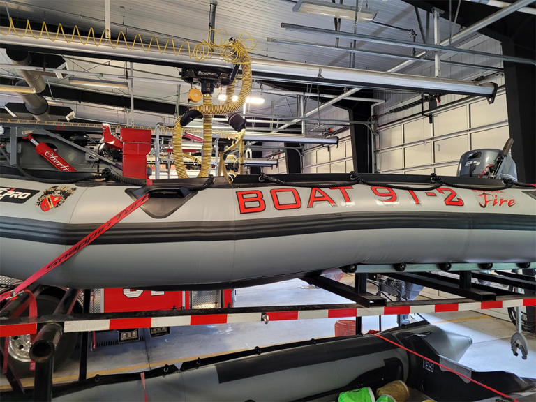 A gray inflatable rescue boat labeled "BOAT 91-2 Fire" is mounted on a trailer inside a fire station. It is secured with red straps, and various equipment is visible around it. The setting is a large garage with overhead lighting.