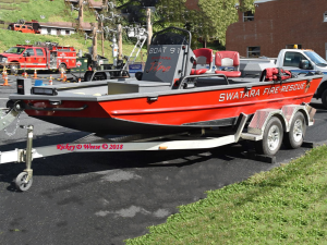 A red Swatara Fire-Rescue boat sits on a trailer, parked on the street. In the background, there are emergency vehicles and equipment.