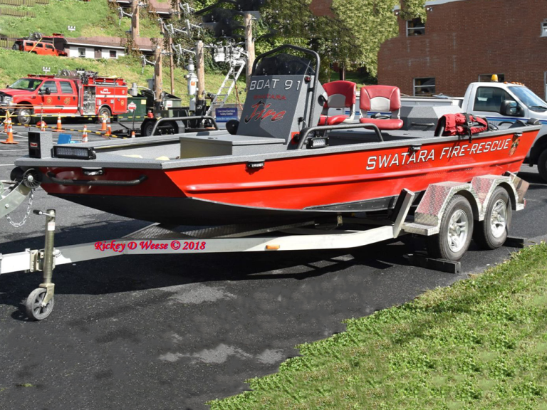 A red Swatara Fire-Rescue boat sits on a trailer, parked on the street. In the background, there are emergency vehicles and equipment.