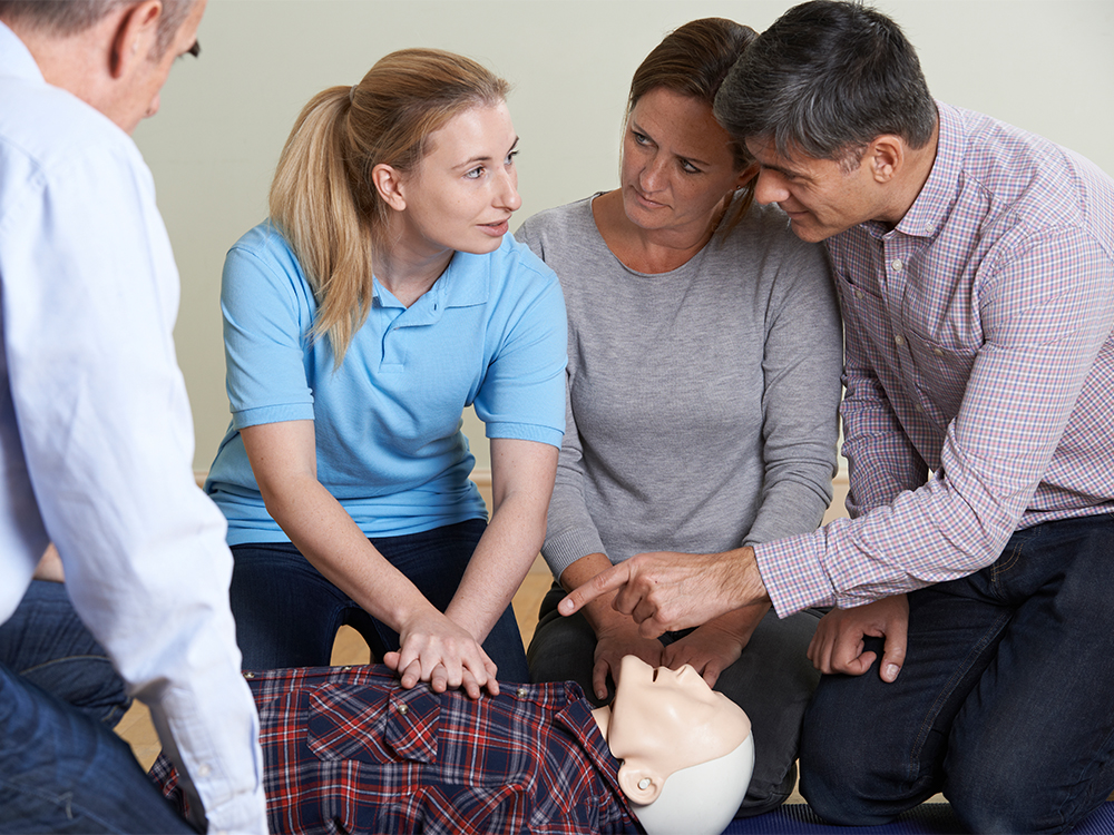 Four people are gathered around a CPR training mannequin. A woman in a blue shirt is demonstrating chest compressions, while the others watch attentively. One person is pointing, possibly giving instructions or feedback.