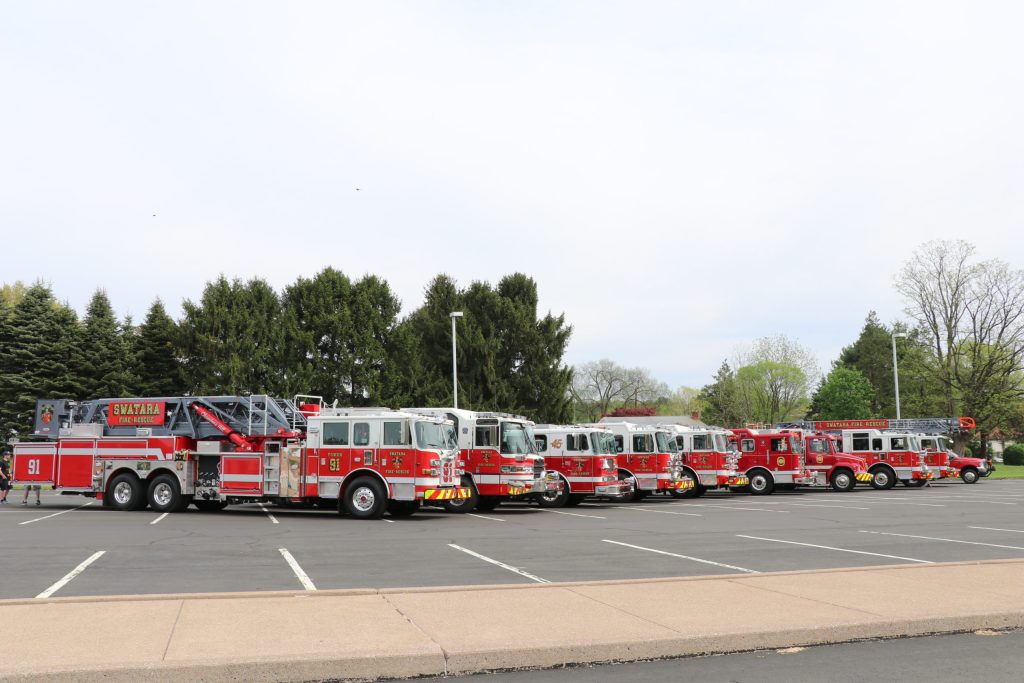 A lineup of seven red fire trucks is parked in an empty lot. The trucks have ladders and various equipment. In the background, there are tall green trees and a cloudy sky.