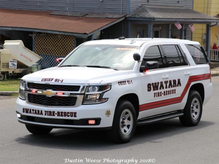 A white Chevrolet SUV marked "SWATARA FIRE-RESCUE" and "CHIEF 91-1" is parked on a street. It has a red and black stripe design and emergency lights on the front. A building with American flags is in the background.