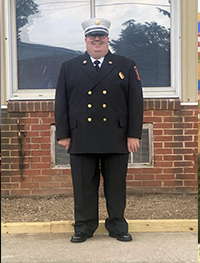 A person in a formal fire department uniform with a white cap stands in front of a brick building with two windows.