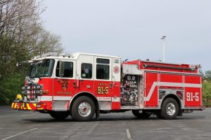 A red and white fire engine parked on an asphalt surface, surrounded by trees. The truck displays the number 91-5 and various equipment compartments. The sky is overcast.