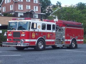 A red and white fire truck is parked on a road in front of a multi-story house with a pointed roof. The fire truck has multiple compartments and hoses, with the number 45 and fire department insignia visible. Trees are in the background.