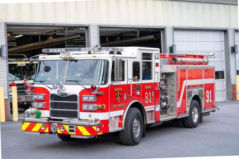 A large red and white fire truck with "Engine 91" labeled on its side is parked outside a garage bay. The vehicle features multiple compartments and equipment for firefighting, with reflective safety markings on its front bumper.