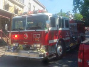A red fire truck with the number 91 displayed on the side is parked on a street. The truck has various equipment attached, and residential buildings are visible in the background.