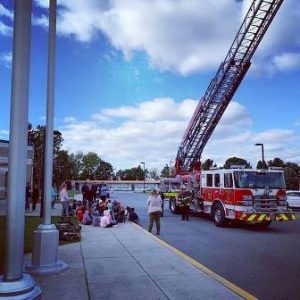 A fire truck with a raised ladder is parked on a road under a partly cloudy sky. A group of people, including children, are gathered on the sidewalk, some sitting and watching the scene. Trees and a building are in the background.