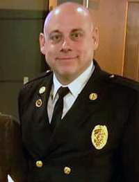 A person in a formal dark uniform with a badge and buttons stands indoors, smiling. They have a bald head and are wearing a white shirt with a black tie under the uniform. The background features wooden paneling and a partial view of another person.