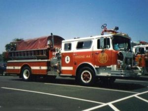 A red and white fire truck, labeled "Engine 44," is parked on a paved lot under a clear blue sky. The truck features a side hose and ladder, with a covered back section.