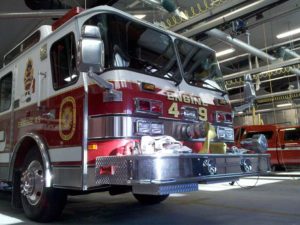 A red and white fire truck, labeled "Engine 49," is parked inside a fire station. The truck has chrome elements and various equipment visible. Fluorescent lights and hoses hang from the station's ceiling.