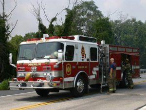 A red and white fire truck with "Oberlin" and "Engine 43" on its side is parked on a road. A firefighter in uniform is standing next to it, preparing equipment. Trees and greenery are visible in the background.
