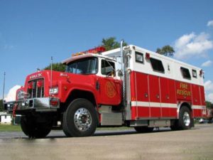 A red and white fire rescue truck is parked on a pavement under a clear blue sky. The vehicle is marked with "Fire Rescue" and features a shiny chrome front with red signal lights and equipment storage compartments. Trees are visible in the background.