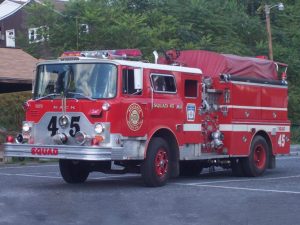 A red fire truck labeled "Squad 45" is parked on a road. It features hoses, a water tank, and emergency lights. Trees and a building are visible in the background.