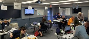 A busy emergency operations center with people working on laptops. Signs overhead indicate different roles like "Fire Service Liaison" and "Radiological Officer." A large screen displays information, and maps are visible on the walls.