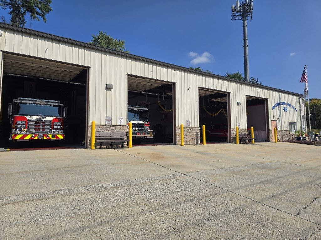 A fire station with open garage bays, revealing fire trucks inside. The building is light-colored with yellow posts at each entrance. An American flag is flying on a pole to the right. The sky is clear and blue.