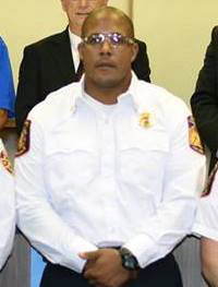 A person in a white uniform shirt with embroidered patches, standing with hands clasped in front. The individual is wearing glasses and appears to be part of a formal group photo with others partially visible.