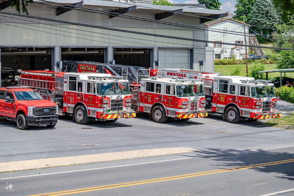 Three red fire trucks and a red pickup truck are parked in front of a fire station. The vehicles are aligned in a row on a sunny day, with a residential neighborhood visible in the background.