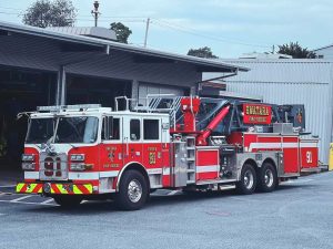 A red and white fire truck labeled "Swatara" and "91" is parked outside a fire station. The truck features multiple compartments and a ladder on top. The background shows an open garage and surrounding trees.
