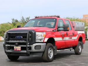 A red fire department utility truck is parked in an outdoor area. The truck has a Ford logo on the front, emergency lights on top, and is marked with "Fire" and "Utility 456." There's a storage compartment on the back. Trees and a building are in the background.