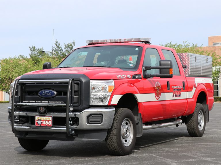 A red fire department utility truck is parked in an outdoor area. The truck has a Ford logo on the front, emergency lights on top, and is marked with "Fire" and "Utility 456." There's a storage compartment on the back. Trees and a building are in the background.