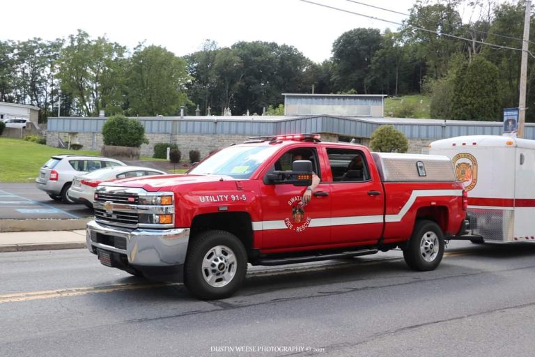 A red utility fire department truck with "Utility 91-5" marked on the side is pulling a trailer. It is driving on a road, with parked cars and trees in the background.