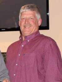 A man with short gray hair and a mustache is smiling, wearing a long-sleeved maroon shirt. The background shows a wall with a dark, flat object, possibly a TV or picture frame.