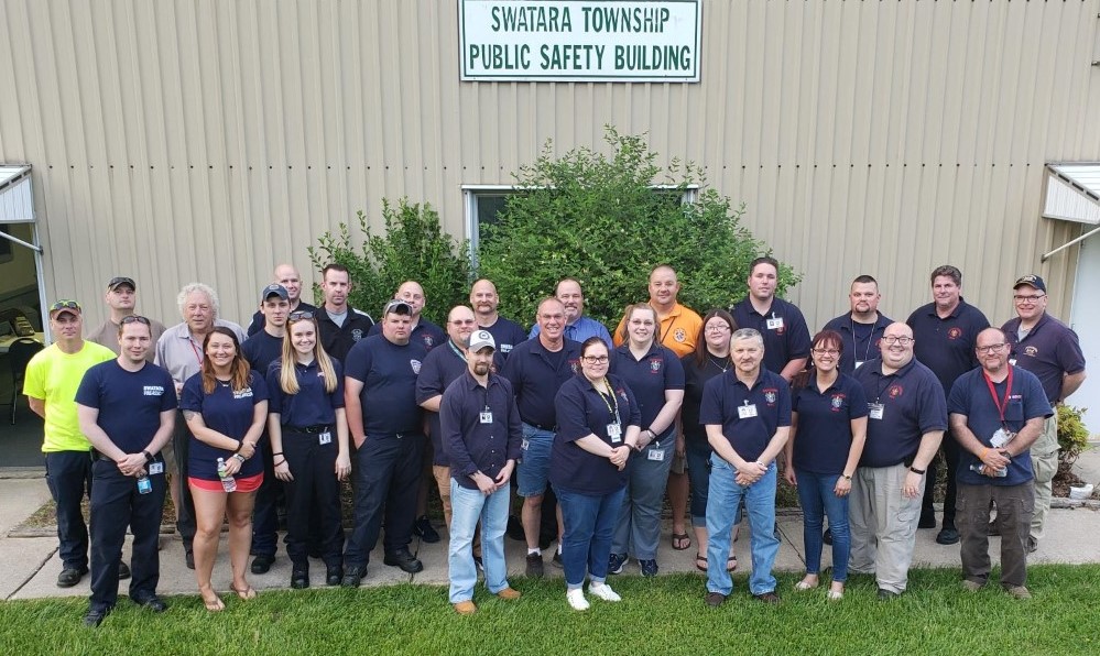 A group of people poses in front of the Swatara Township Public Safety Building. They are wearing casual and uniform-style clothing, standing on a lawn with a building and greenery in the background.
