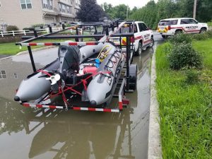 A rescue boat on a trailer is parked on a flooded street beside a white SUV labeled "Swatara Township." Another SUV is nearby, with water covering the road. Residential buildings and greenery are in the background.