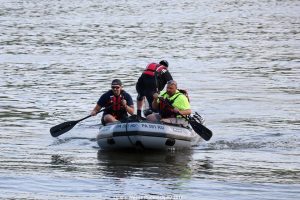 Three people in life jackets are on a small inflatable boat on a body of water. Two are seated, holding paddles, while the third is standing and adjusting something at the rear. The setting appears calm, with gentle water ripples.