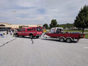 A red pickup truck with a camper shell is parked in a parking lot, towing a red and silver boat on a trailer. The background shows a JCPenney store and scattered trees under a clear sky.