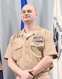 A person in a tan military uniform stands in front of flags, with arms at their sides. They have short hair and various badges and insignia on the uniform.