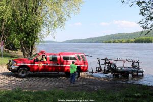 A red fire department truck is parked on a boat ramp by a river. Two individuals are loading or unloading a small boat on a trailer. Lush greenery and a hill are visible across the river under a clear blue sky.