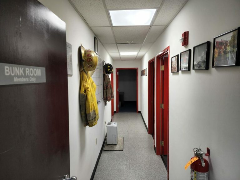 A hallway in a fire station with white walls and a gray floor. A yellow firefighter jacket and helmet hang on hooks. A sign on the door reads "Bunk Room Members Only." Pictures line the wall, and a fire extinguisher is on the floor.