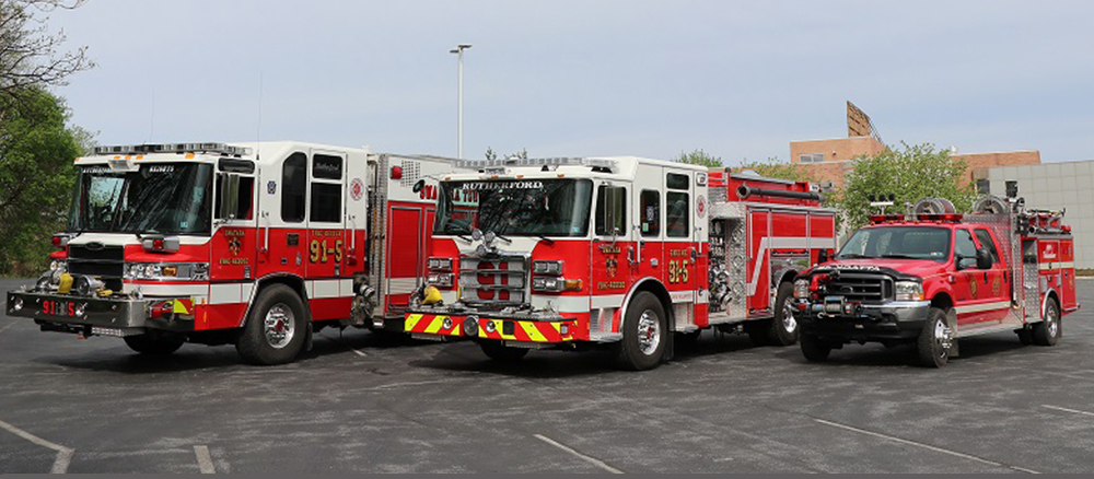 Three red and white fire trucks are parked side by side in a parking lot. The trucks have various equipment and number markings. Trees and a building are visible in the background.