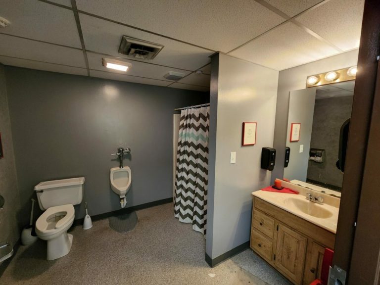 A bathroom with a toilet, urinal, and shower with a chevron-patterned curtain. There is a sink with a mirror above, a soap dispenser, and a red object on the counter. The walls are painted gray, and the floor has a speckled appearance.