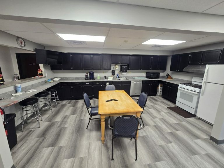 A kitchen with dark cabinets, a light wood table surrounded by blue chairs, and stainless steel appliances, including a stove, fridge, and dishwasher. The floor has gray wood-patterned tiles, and a wall-mounted TV is visible.