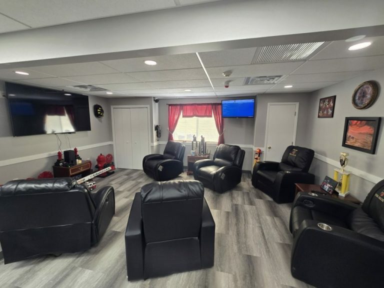A cozy room with several black reclining chairs arranged around the space. The walls have a clock, paintings, and two flat-screen TVs. A window with red curtains provides natural light. Various decorations and a trophy are also visible.