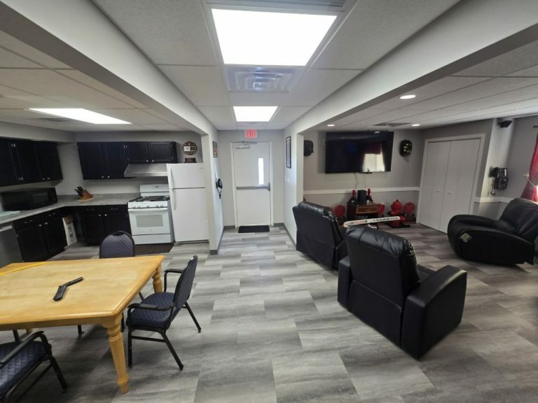 A spacious living area featuring a kitchenette with black cabinets, a white refrigerator, and stove. There's a wooden dining table with chairs in the foreground and a comfortable seating area with recliners and a large wall-mounted TV in the background.