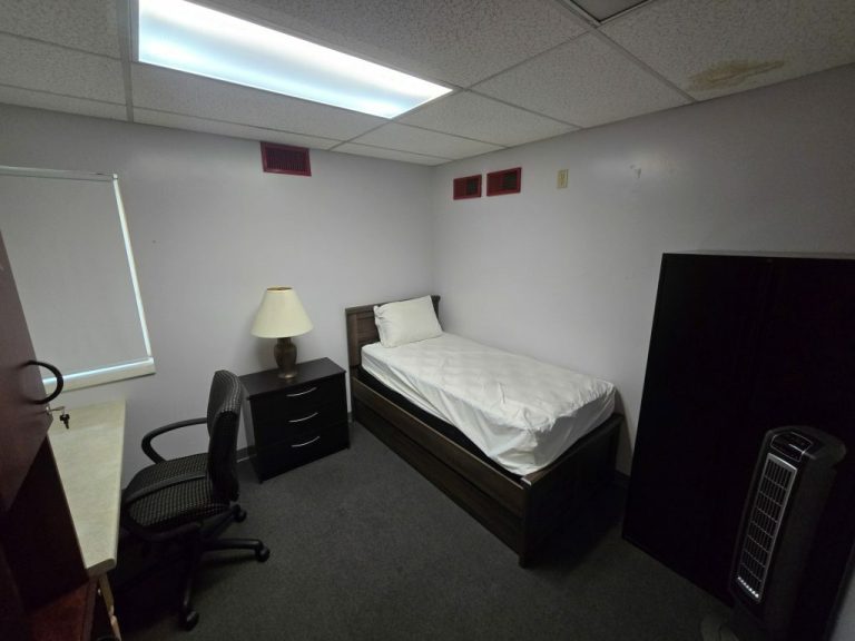 A small, dimly lit room with a single bed, white bedsheet, and pillow. A black desk with a lamp is next to the bed. There is an office chair, a cabinet, and a tower fan. The ceiling shows signs of water damage.
