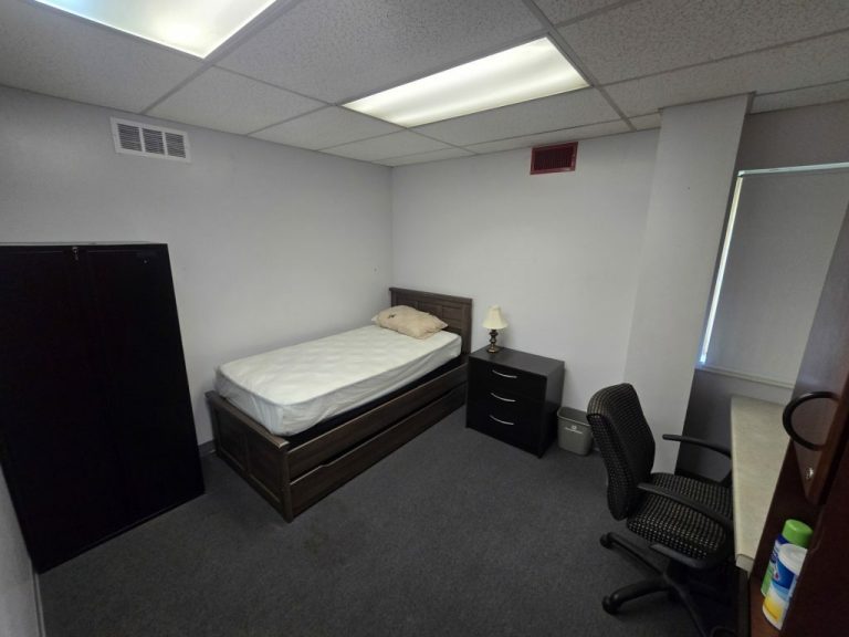 A small, minimalistic room with a twin bed, a pillow, a bedside table with a lamp, a wardrobe, and a desk with a chair. The carpet is gray, and the walls are light-colored. Fluorescent lights illuminate the room.