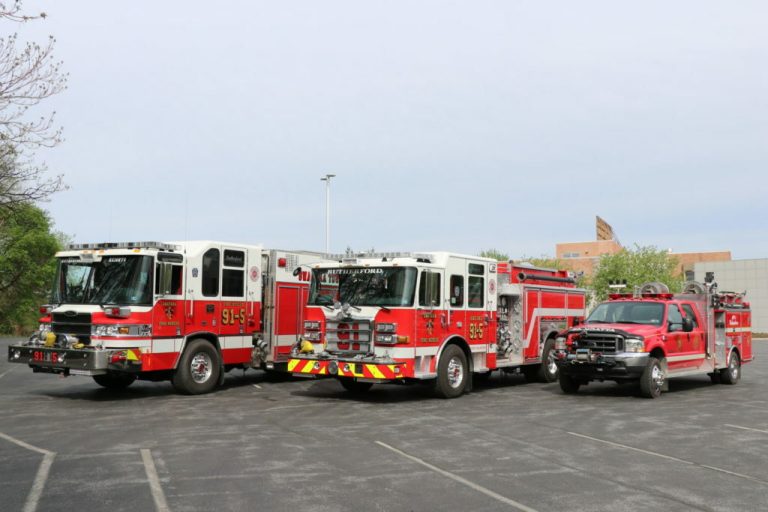 Three fire trucks are parked on a paved lot during the day. Two large fire engines on the left display red and white colors, while a smaller red utility vehicle is on the right. Trees and buildings are visible in the background.