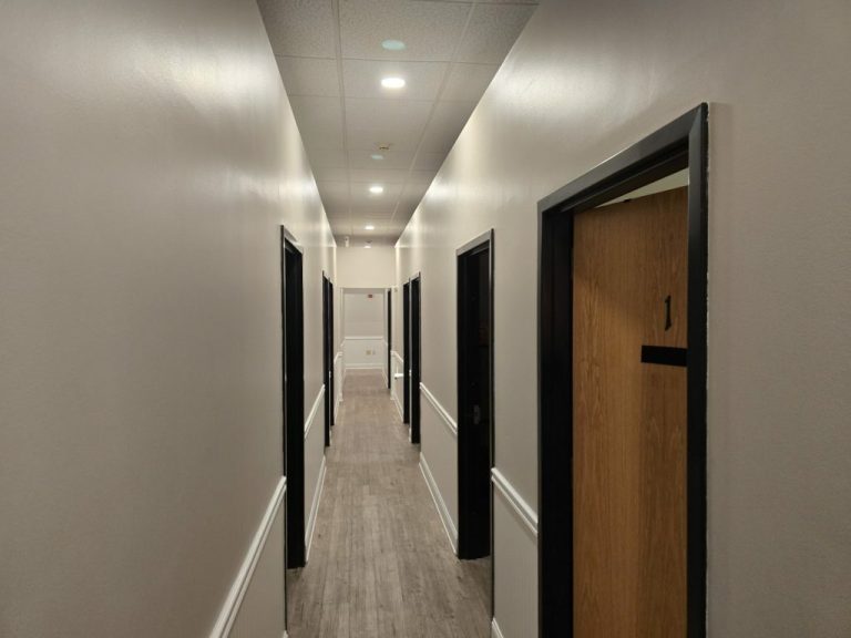 A long, narrow hallway with light beige walls and a wooden floor. Doors with black frames line both sides, each numbered, with "1" visible in the foreground. The ceiling features recessed lighting, creating a clean and modern look.