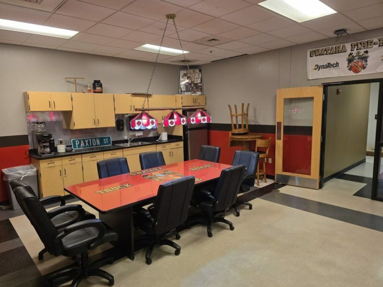 A break room with a rectangular table and eight red and black chairs. The table has a "TRUCK" design. Cabinets line the wall, and a coffee maker sits on the counter. A neon light hangs above, and a "PAXTON ST." street sign is on display.