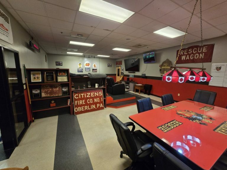 A meeting room with a long table surrounded by chairs. Walls are decorated with fire department memorabilia, including signs and plaques. A TV is mounted on the wall, and a pool table light fixture hangs above the table.