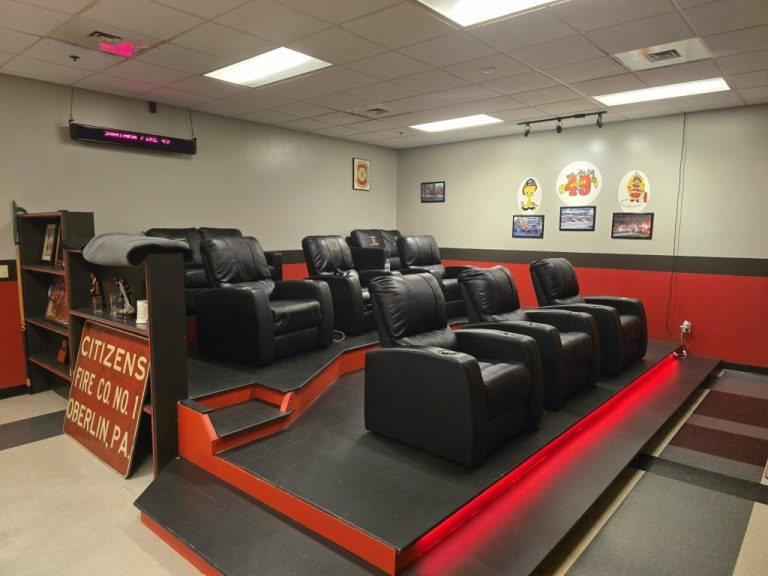 A cozy room featuring black reclining chairs on a raised platform, with walls adorned with various photos. A sign reads "Citizens Fire Co. No. 1, Oberlin, PA." Red and black color theme with overhead lighting completes the ambiance.