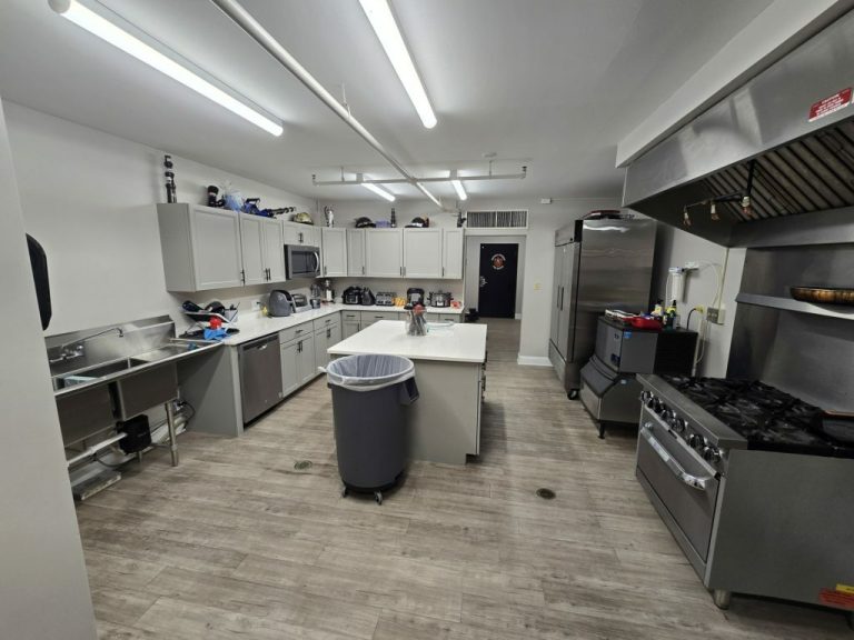 A spacious, modern kitchen with stainless steel appliances, including a large stove and refrigerator. The room features white cabinets, a central island with a sink, and a trash bin. The flooring is light wood, and the lighting is bright and even.